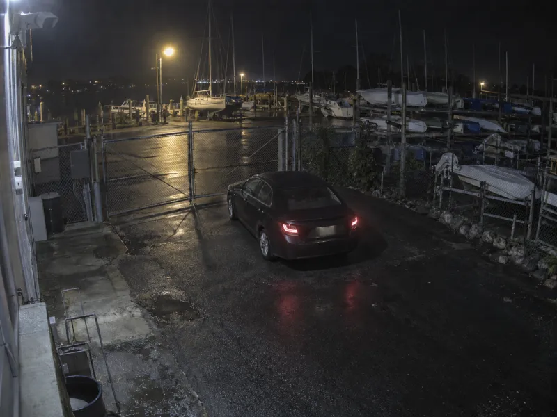 Night camera still of a dark sedan stopped at a closed boatyard gate; no person or plate is readable